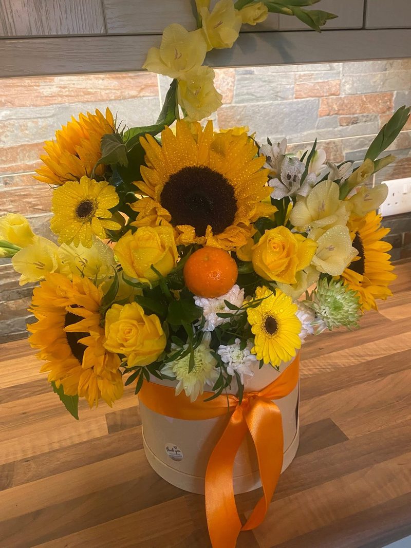 Sunflowers, yellow roses and gladioli in a cream hat box with orange ribbon